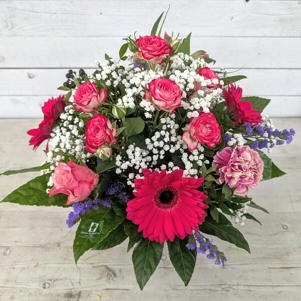 Handgebundener Blumenstrauß mit pinken Gerbera, rosa Rosen, weißem Schleierkraut und violetten Akzenten auf hellem Holzhintergrund.