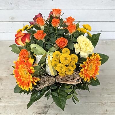 Farbenfroher Blumenstrauß mit orangefarbenen Rosen, Gerbera und gelben Chrysanthemen, handgebunden mit grüner Schleife und Naturdekoration auf Holztisch