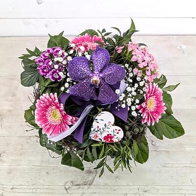 Farbenfroher Blumenstrauß in Violett und Pink mit Vanda-Orchidee, Gerbera, Herz-Deko und Schleierkraut in einem natürlichen Nest auf hellem Holzhintergrund.