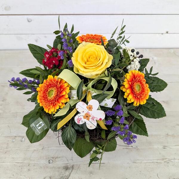 Farbenfroher Gruß Farbenfroher Blumenstrauß in einem Nest mit gelber Rose, orangefarbenen Gerbera, violetten Statice-Blüten und einer grünen Schleife auf hellem Holzgrund.