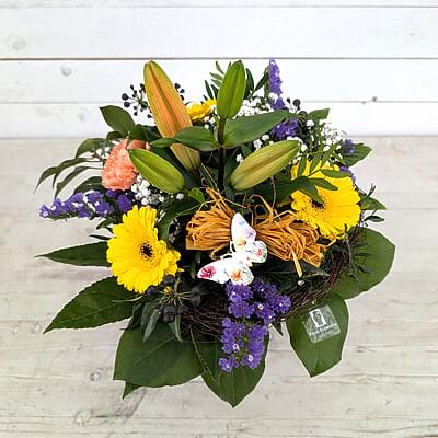 Farbenfroher Blumenstrauß mit gelben Gerbera, orangefarbenen Lilienknospen, lila Statice und weißem Dekoschmetterling in einem rustikalen Zweig-Nest auf hellem Holzhintergrund.