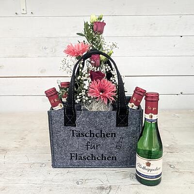 Graue Filztasche mit Aufschrift Täschchen für Fläschchen gefüllt mit pinken Gerbera Rosen und vier kleinen Rotkäppchen Sektflaschen vor weißer Holzwand.