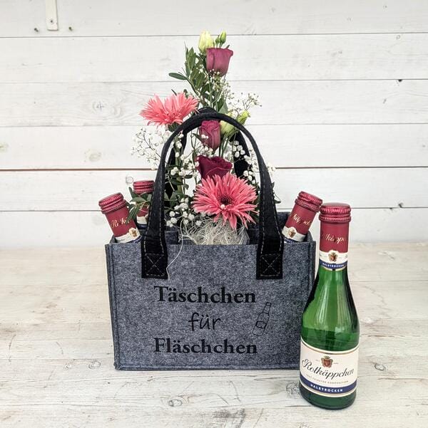 Graue Filztasche mit Aufschrift Täschchen für Fläschchen gefüllt mit pinken Gerbera Rosen und vier kleinen Rotkäppchen Sektflaschen vor weißer Holzwand.